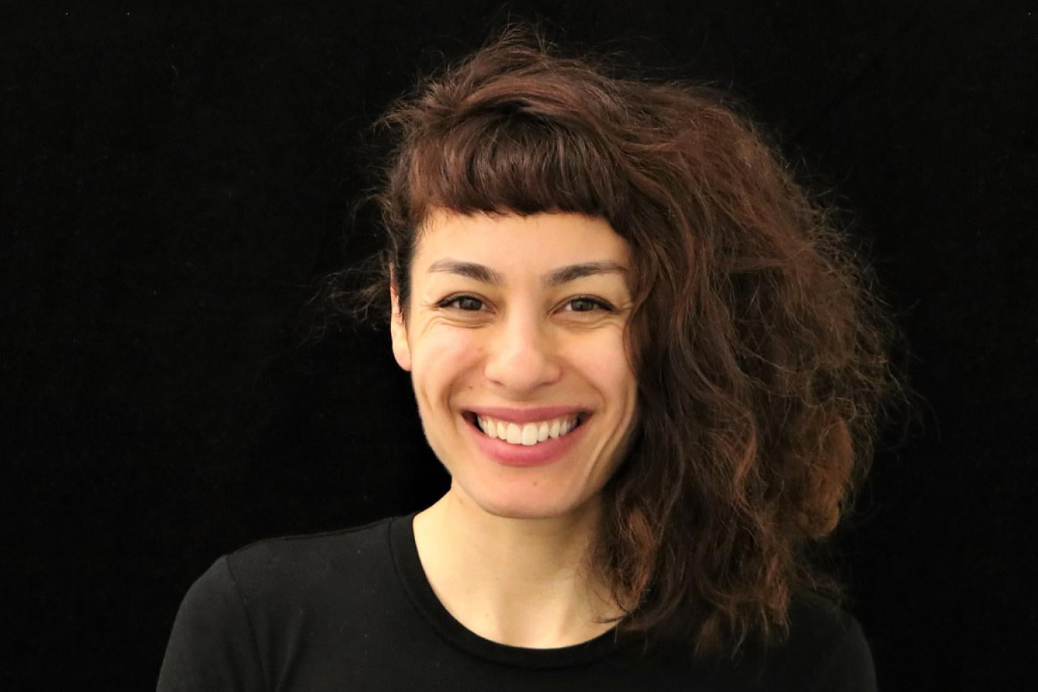 Profile image of a young white woman with shoulder length curly brown hair swept to one side. She is smiling and wearing a black shirt.