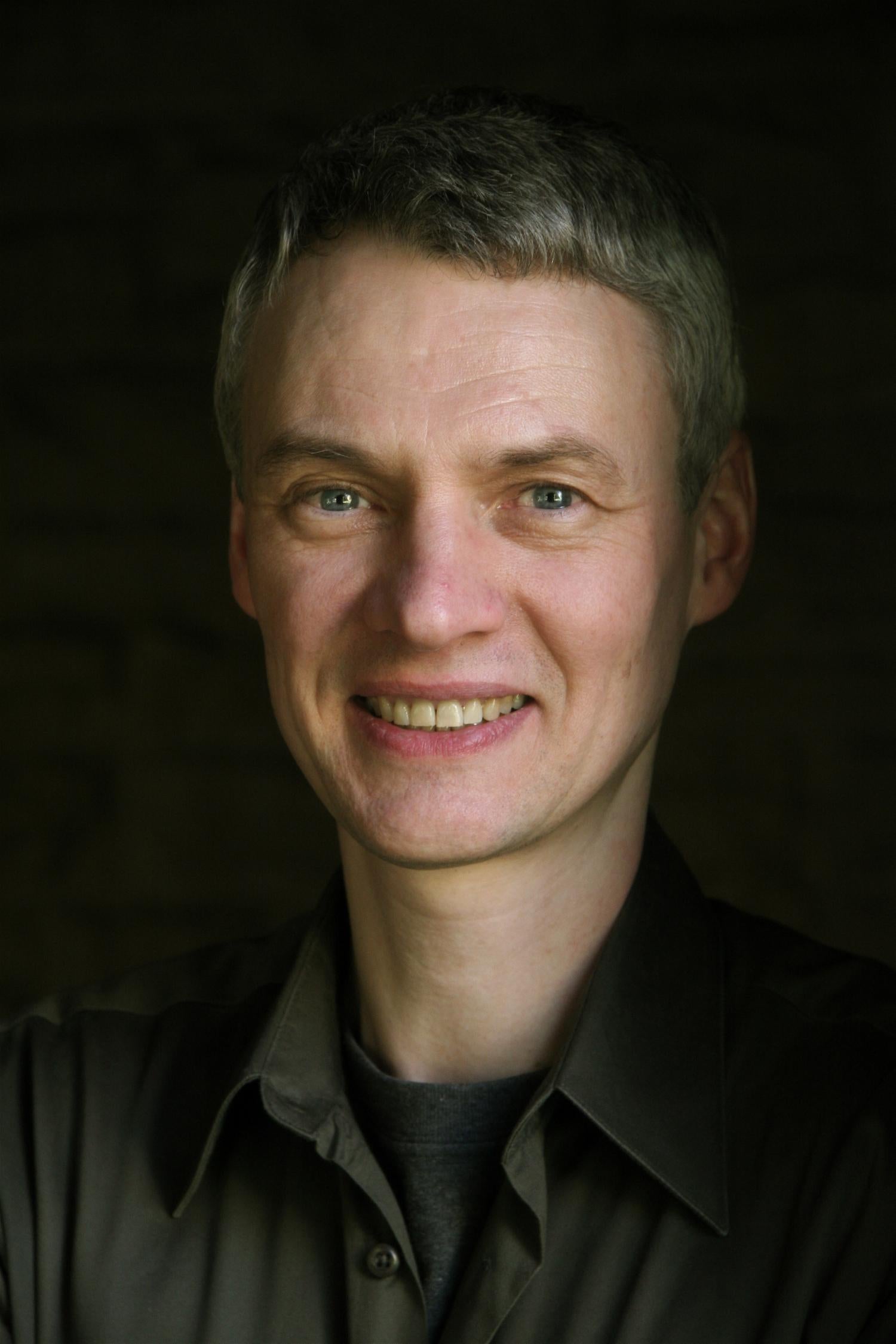 Profile image of an middle-aged white man with short gray hair, smiling. He is wearing a gray button up.