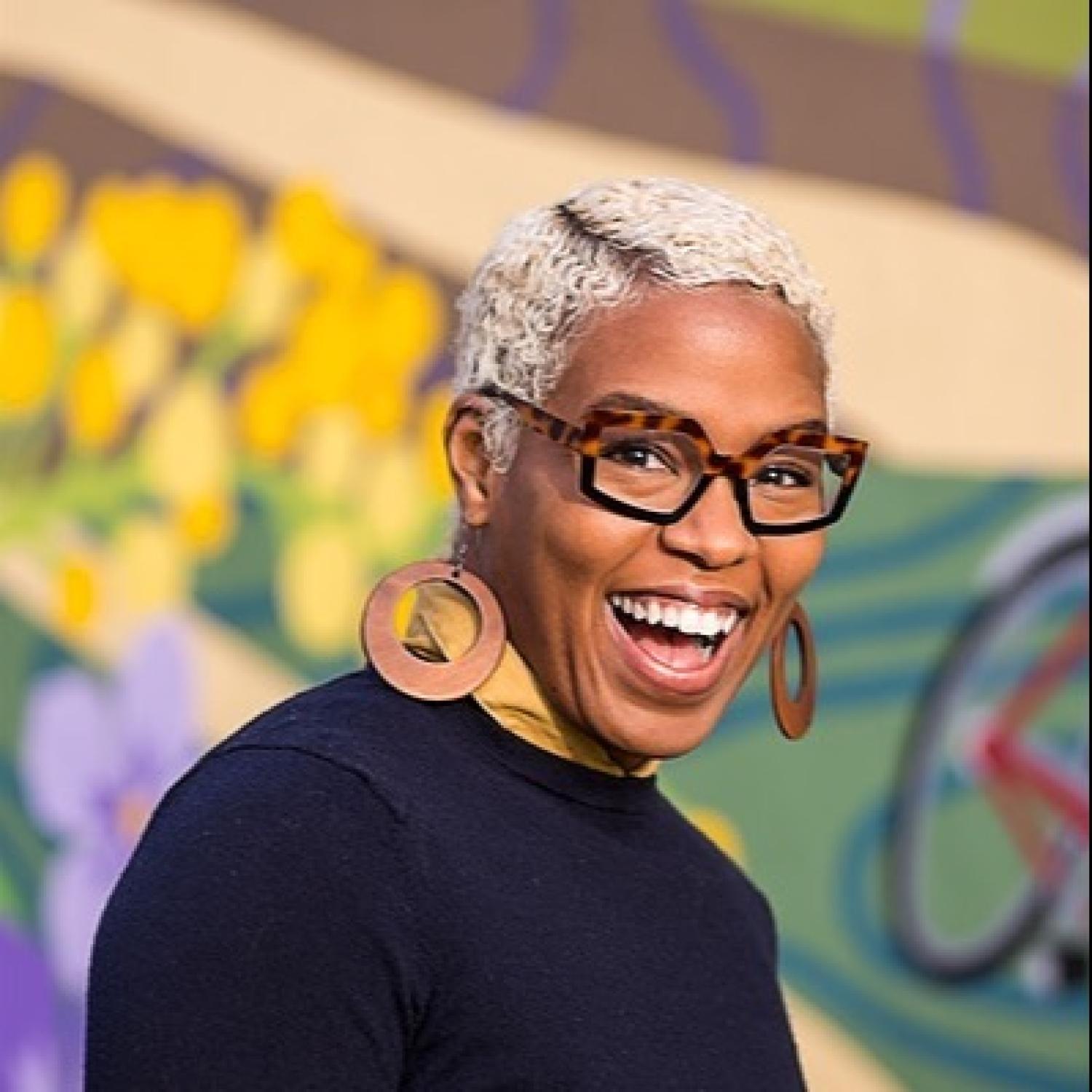 Profile image of a black woman with a white buzzcut, smiling. She is wearing a navy sweater and large wooden earrings.
