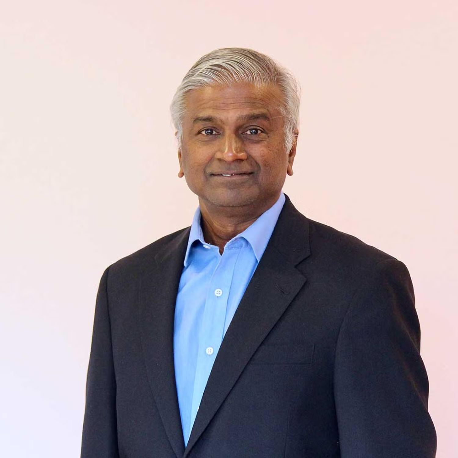 Half-body of a medium brown older man with short gray hair. He is wearing a suit and a blue dress shirt.