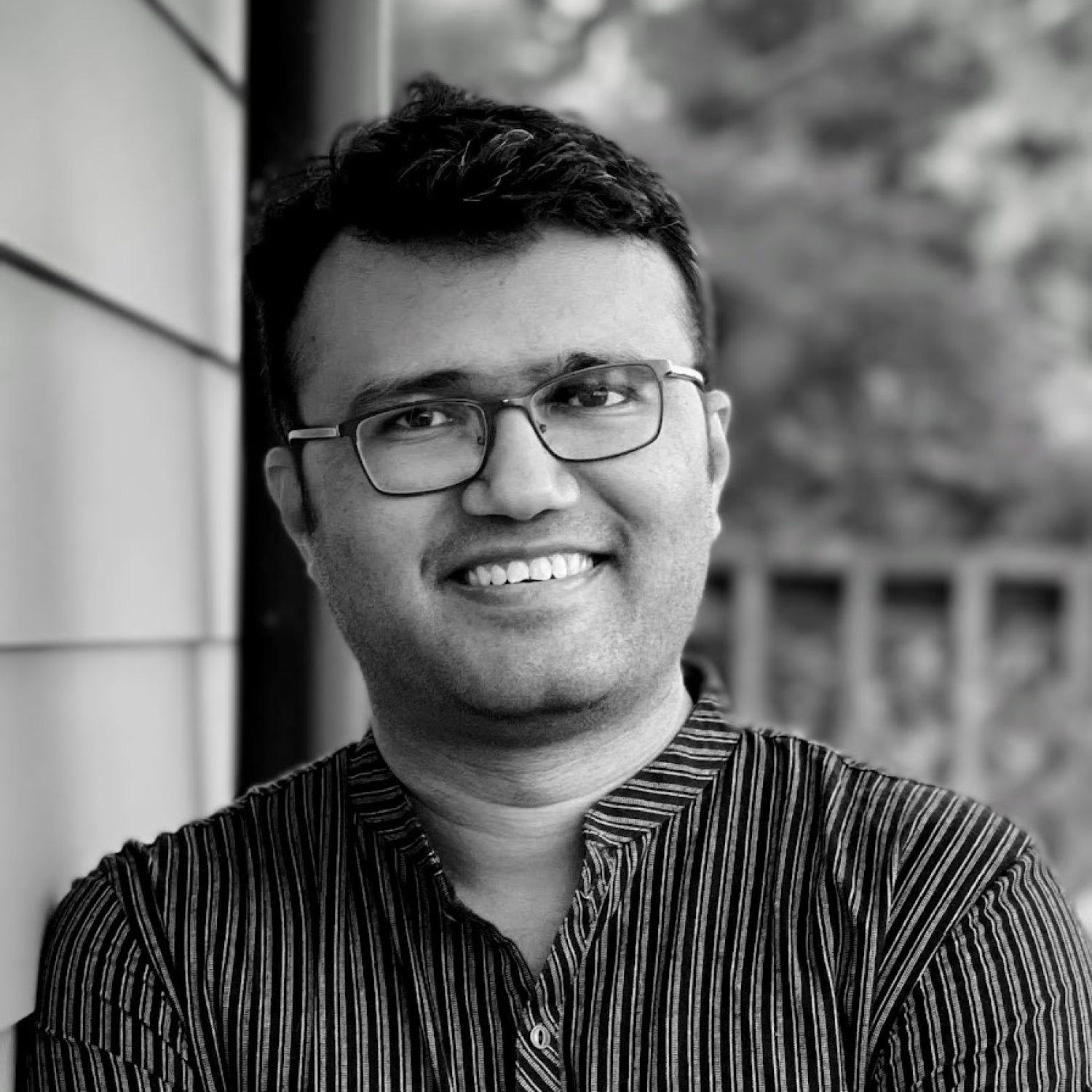 Black and white image of a medium brown man wearing glasses with short black hair, smiling. He is wearing a pinstripe shirt.