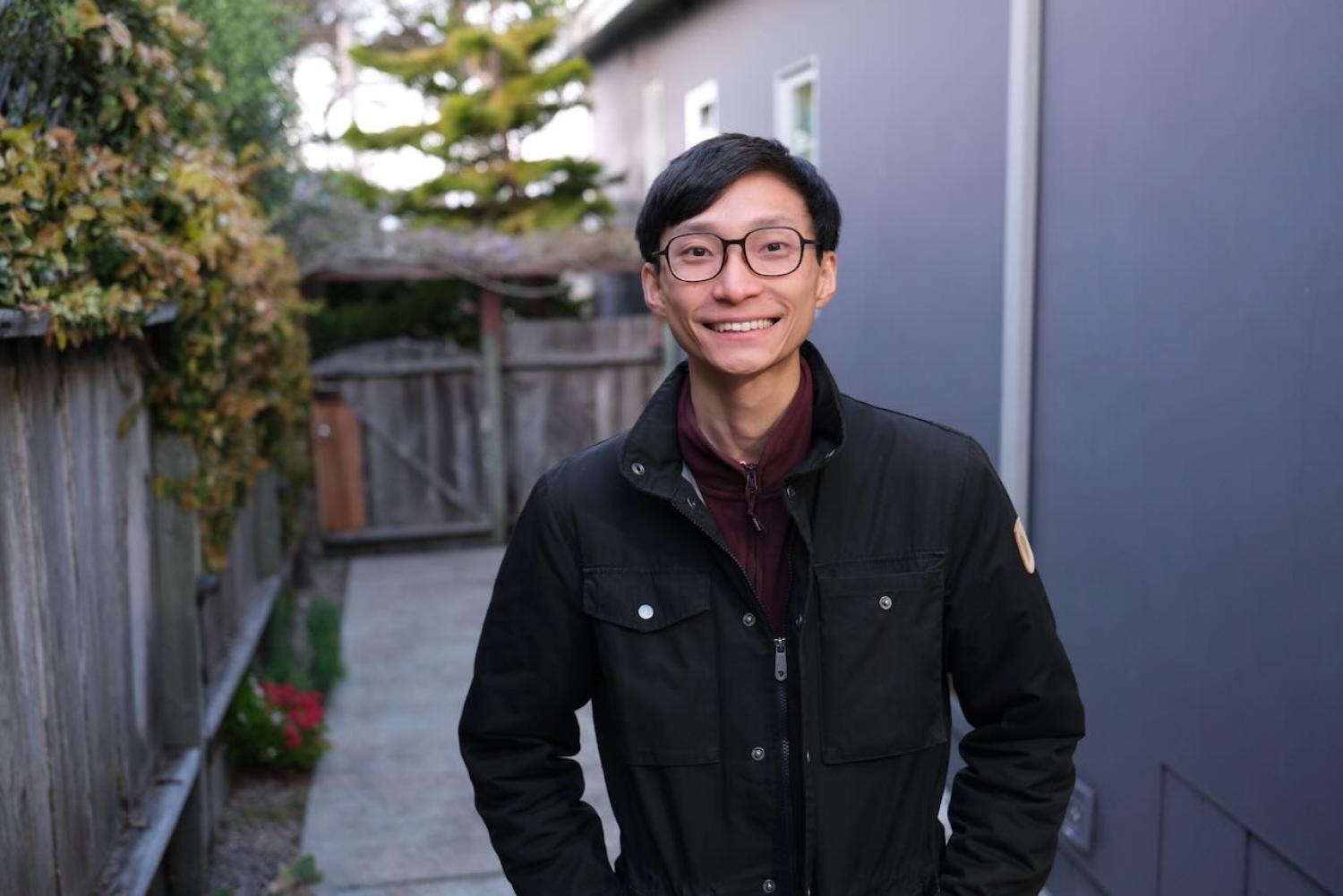 Half-body of a young asian man with short black hair and glasses, smiling. He is wearing a black jacket.
