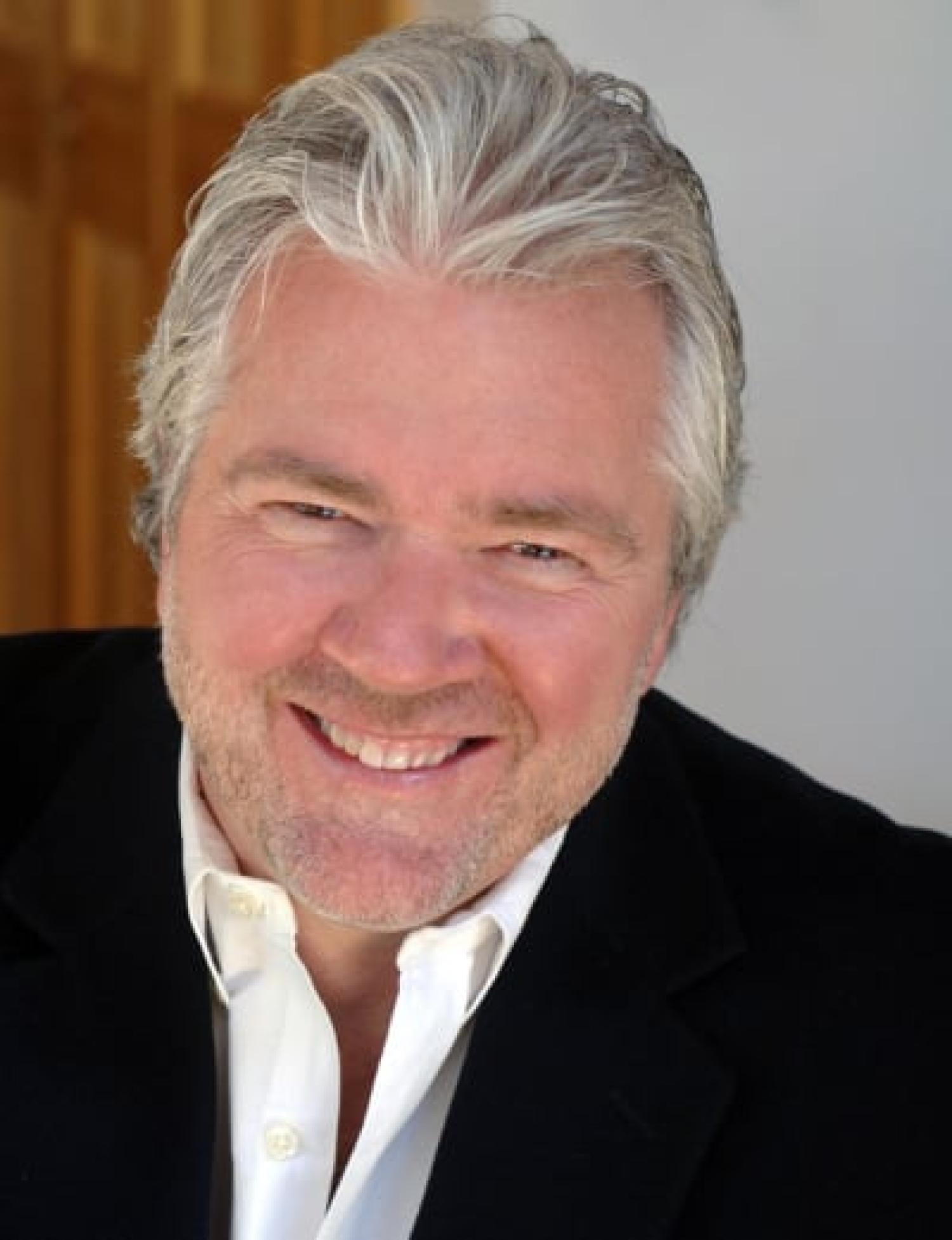 Profile of an older white man with short gray hair and stubble smiling. He is wearing a black suit.