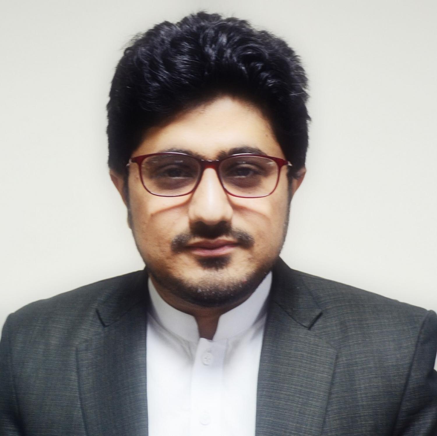 Profile of a brown man with short curly black hair and a beard. He is wearing glasses and a gray suit.