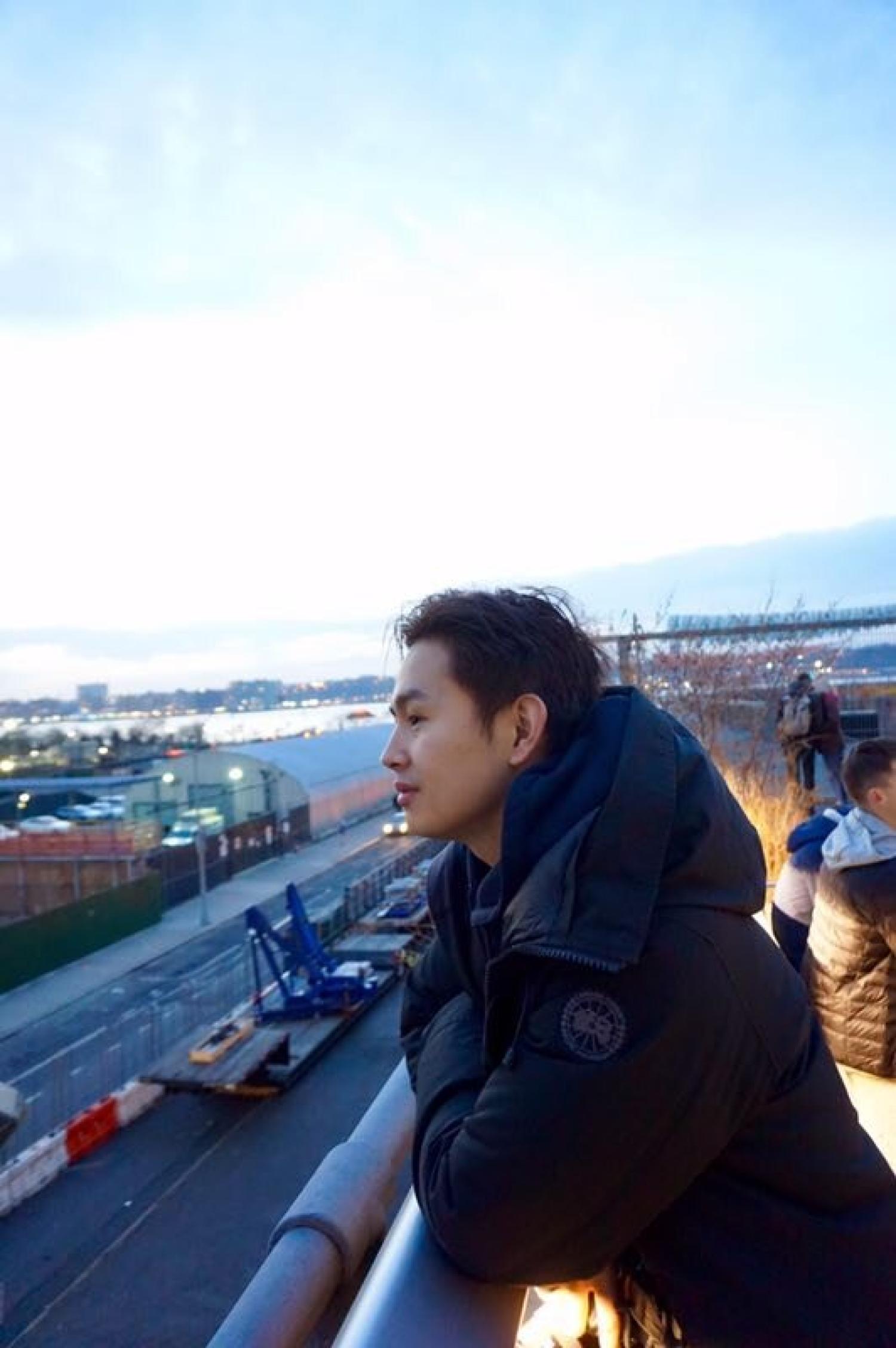 Halfbody of a young asian man leaning on railing overlooking scenery. He has short black hair and is wearing a black puffer jacket.