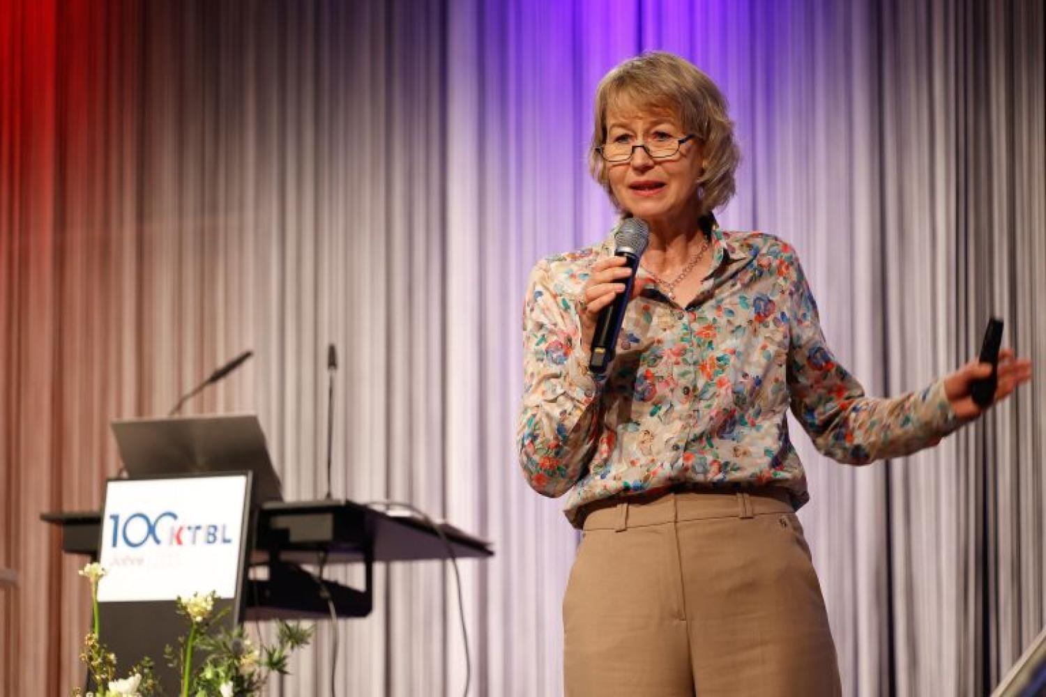 Older white woman talking on stage, holding a microphone.