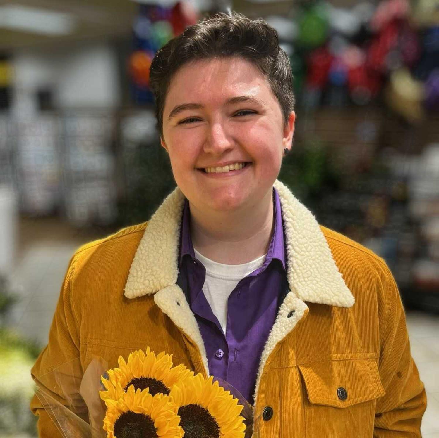 Profile of young white person with short dark hair, holding sunflowers and smiling.