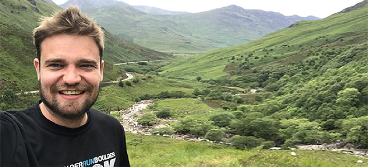 Profile of a young white man standing in front of green mountains.