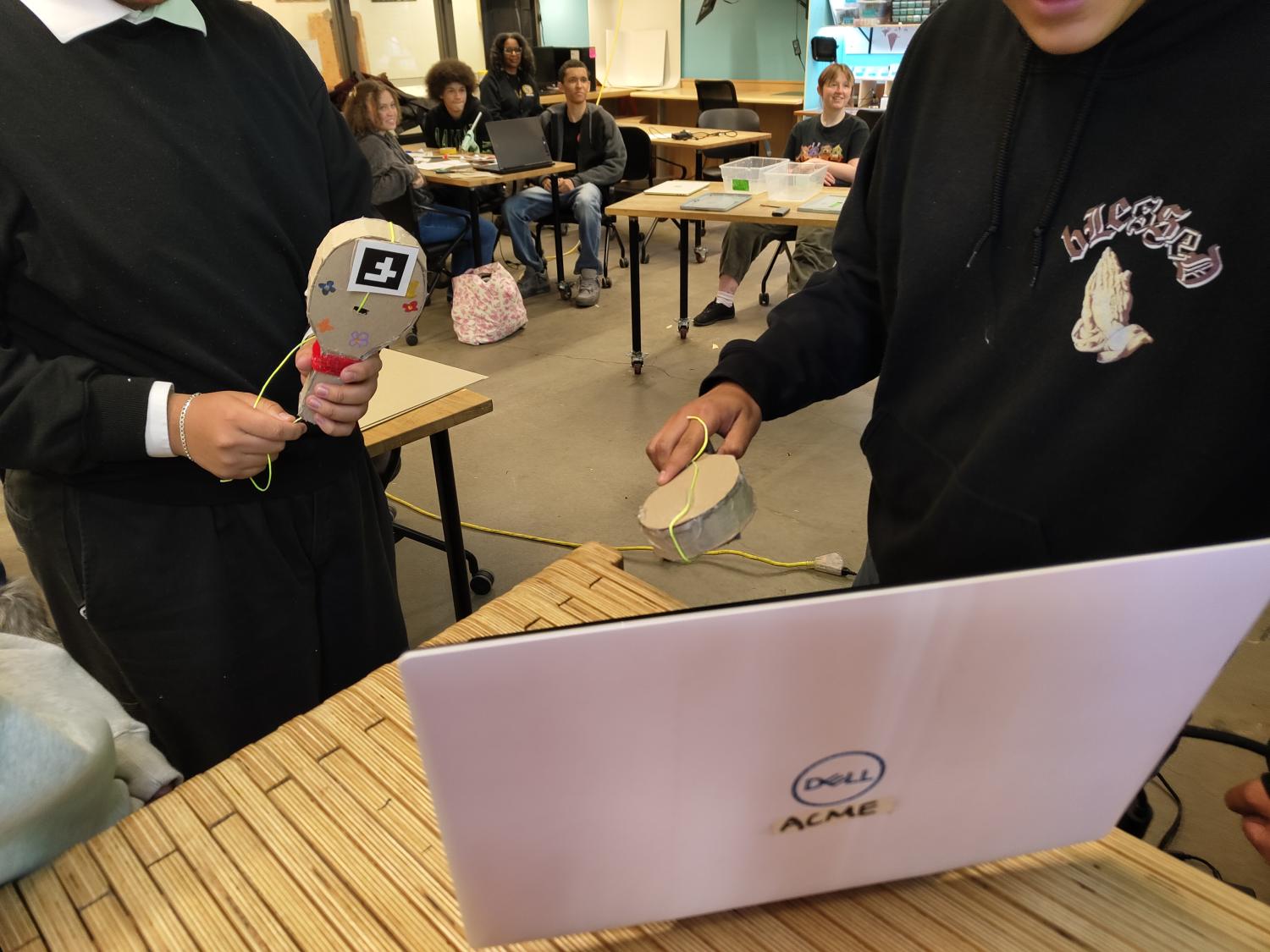 Two people playing with racket shaped cardboard controllers in front of a laptop