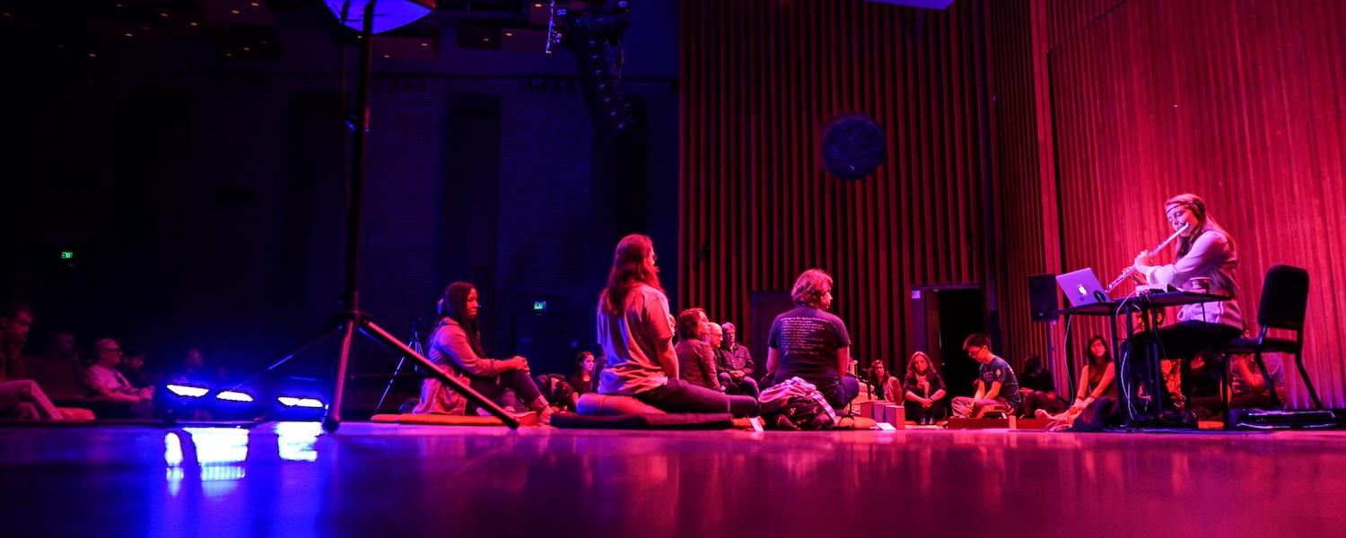 Grace Leslie performs flute with dramatic lighting while audience sits on the floor around her