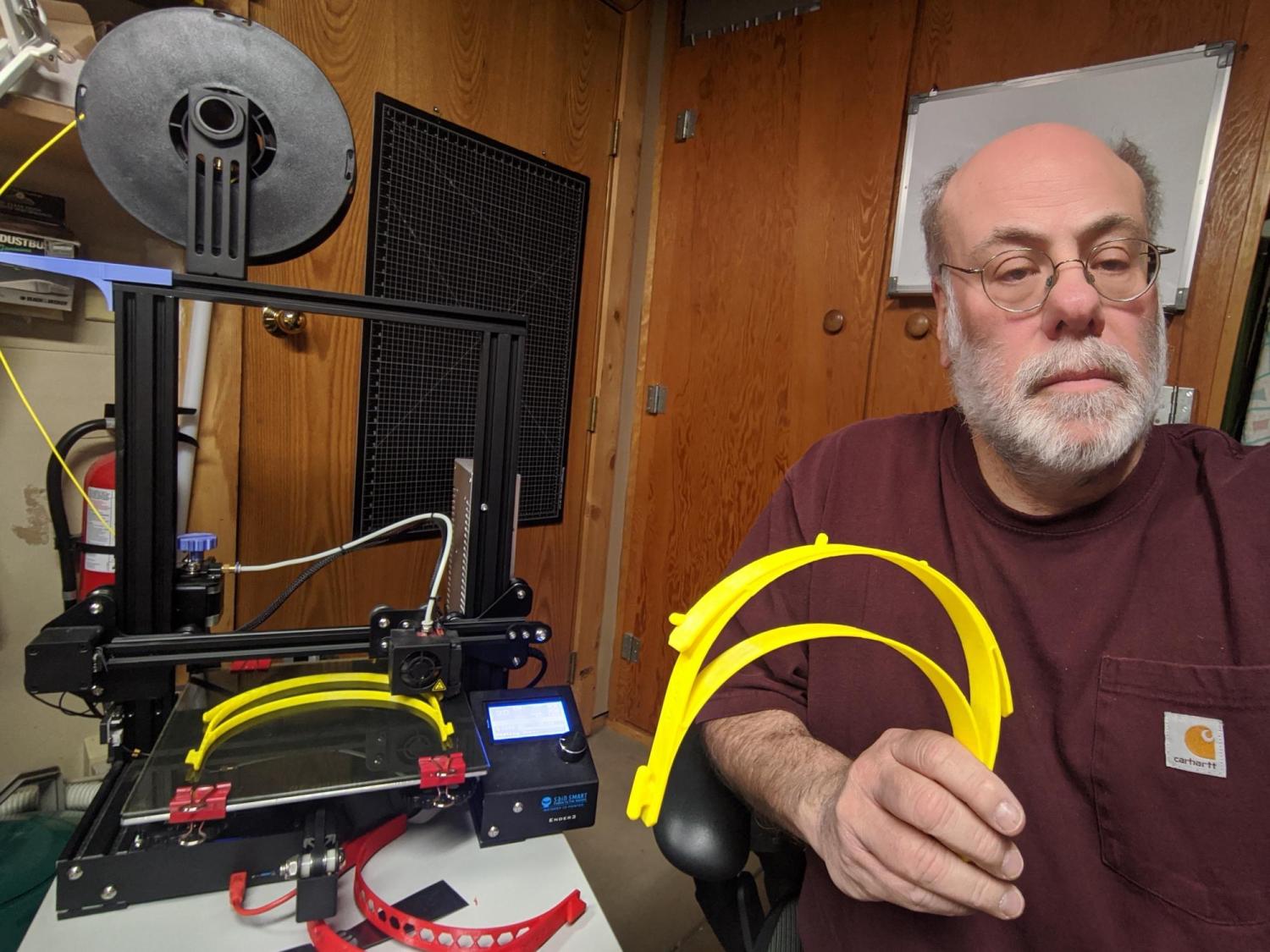 Wayne Seltzer holds several face shield parts next to his 3D printer, which is making several more.