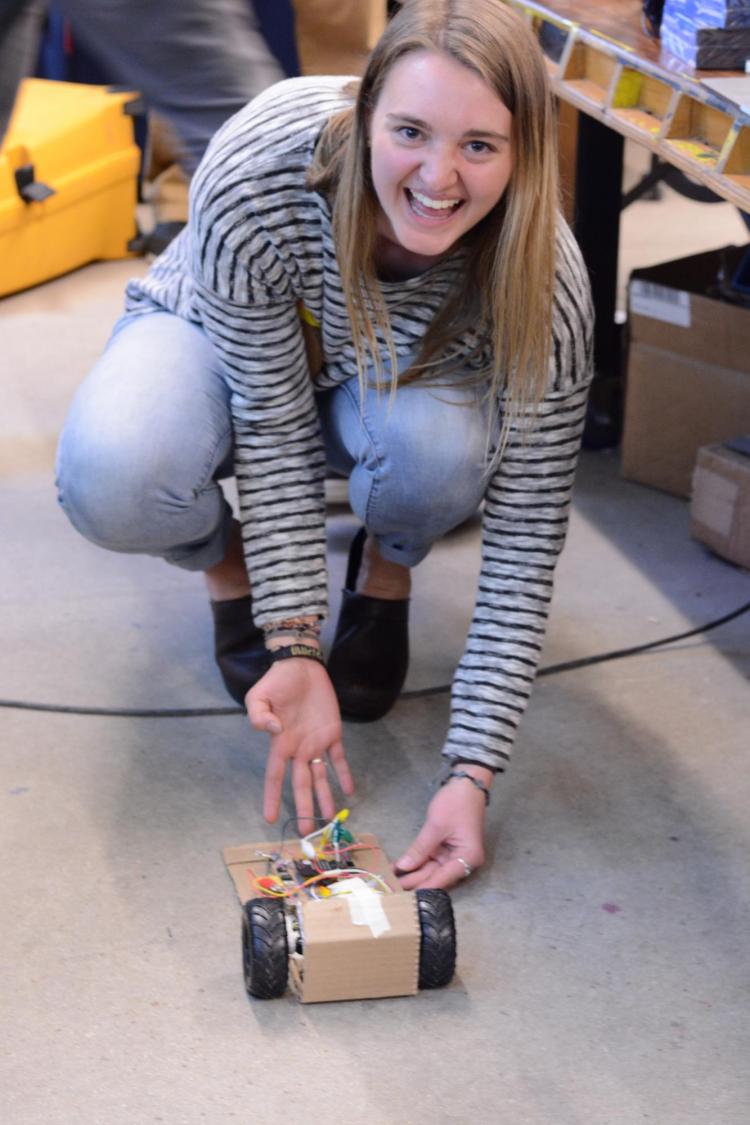 Student crouches on the floor and shows the remote control vehicle she built from a micro:bit, cardboard and remnants from a remote control car.