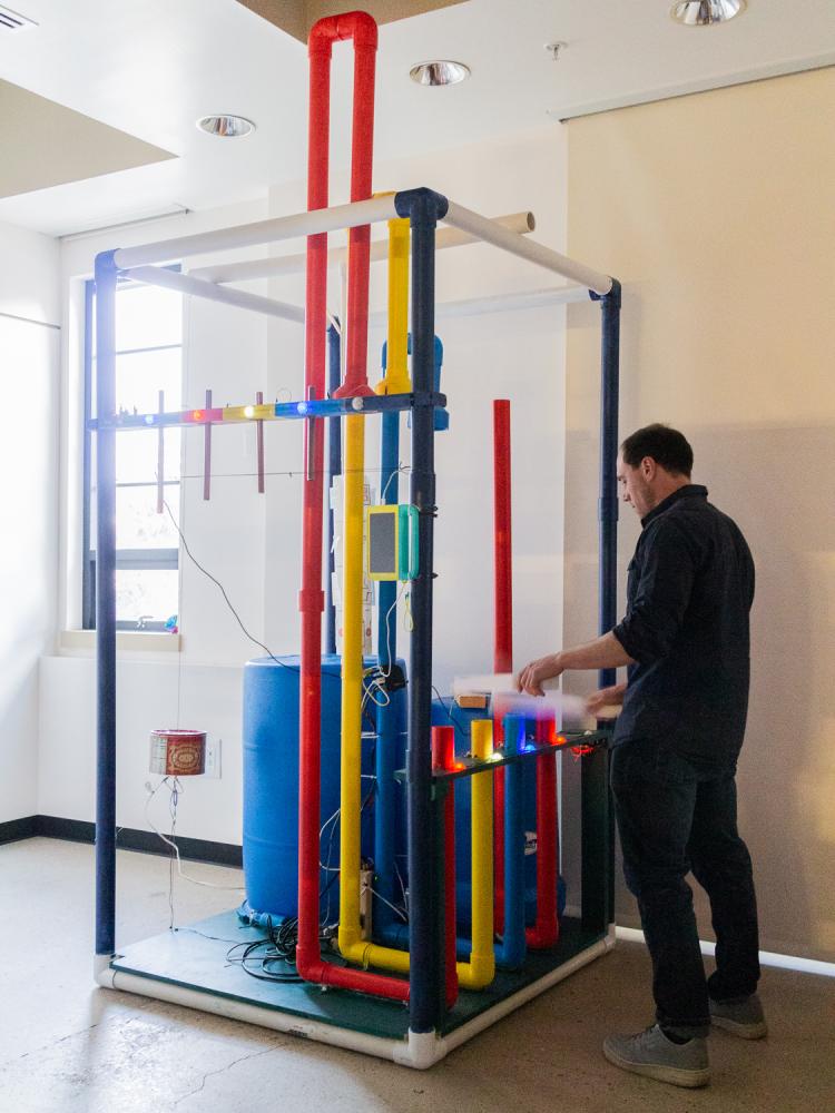 Person stands next to Jam Station, a a 7 feet by 4 feet rectangular PVC cuboid, whereby four instruments crafted from everyday materials are situated so that each member of the jam faces a centrally and vertically oriented neopixel display.