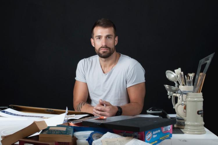 Joel Swanson in a white T-shirt sits behind his desk covered with art supplies.