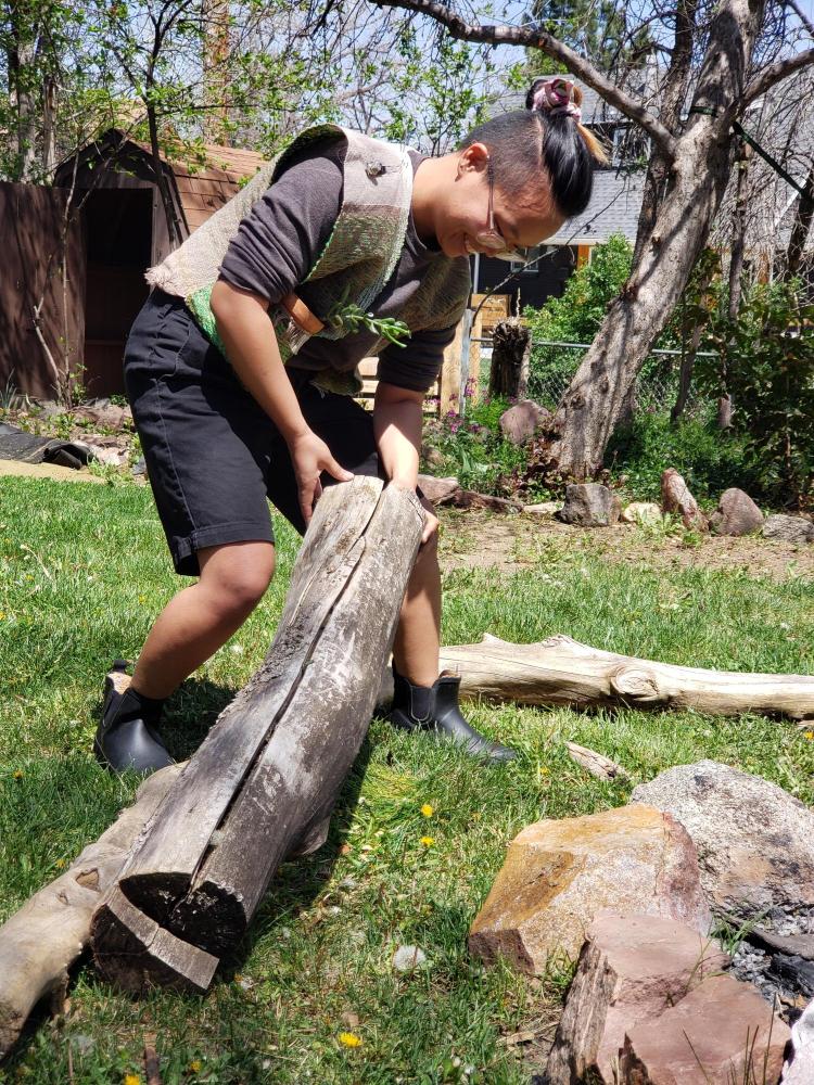 Shanel Wu bends over by a log. Wu wove the vest she is wearing on a traditional 4-shaft floor loom, integrating digital air quality sensors for outdoor workers in polluted environments.