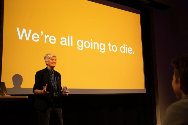 Colleen Macklin smiles in front of screen that says, "We're all going to die."