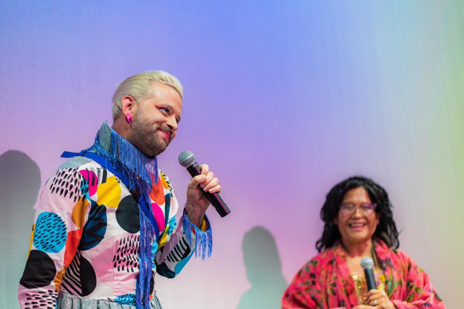 person holding a mic smiling wearing a maud style shirt