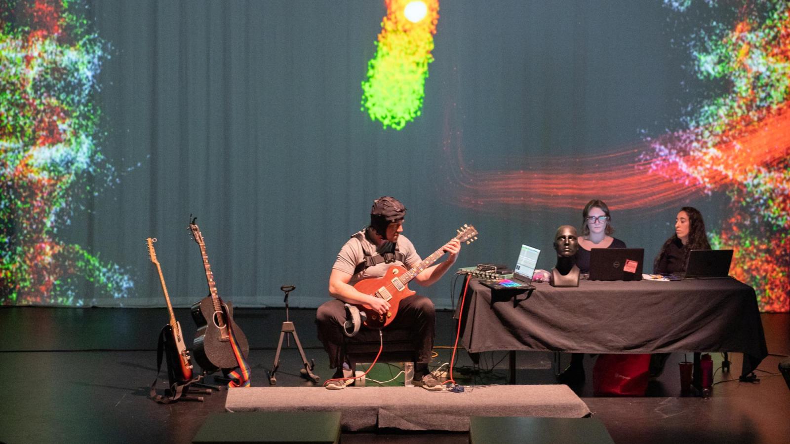 wide shot of man performing on guitar while wearing a brain scanner and projections in the background