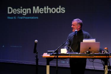 Derek Friday stands behind a podium with a slide projection behind with the words Design Methods