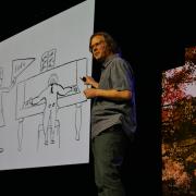 Michael Theodore points to a large cartoon drawing that he drew of a man at a piano and another man holding loud and soft signs.