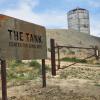 A rusty metal sign that says "The TANK" with the decommissioned water tank on a hilltop in the background