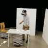Person with VR goggles touches a table that has a Roomba with a mechanical scissor lift below. 