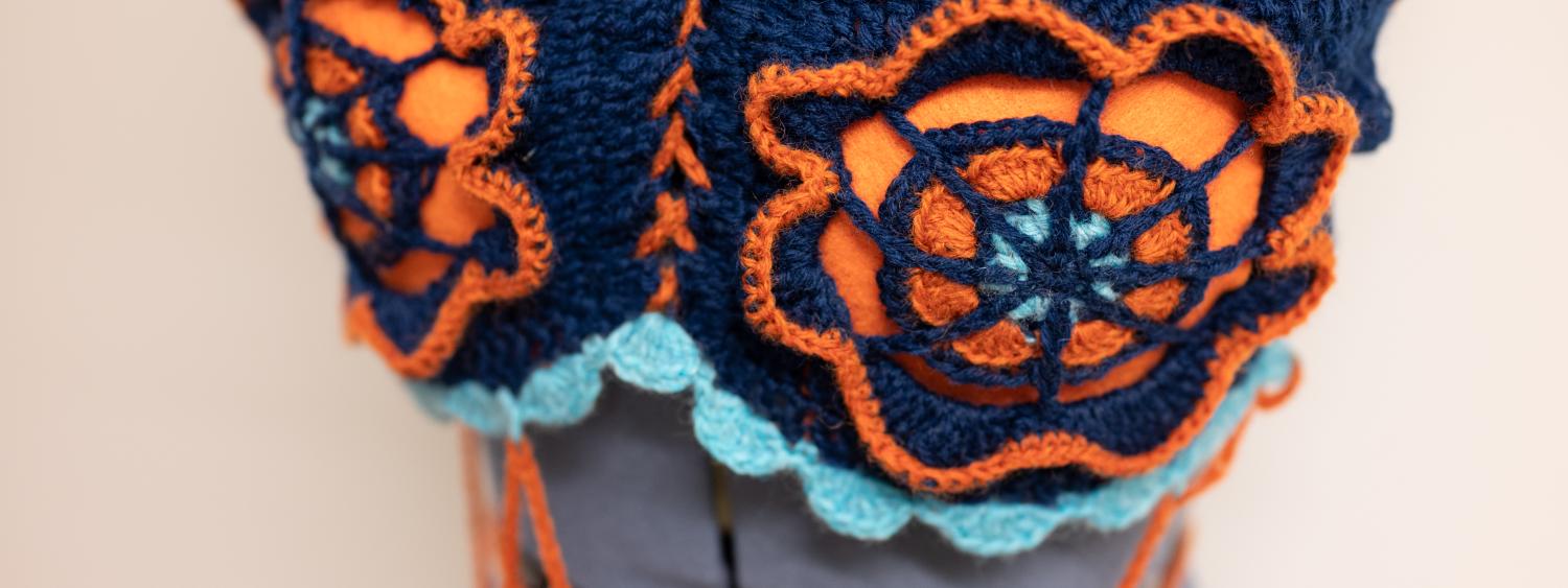 A orange and blue crocheted bralette with a flower designs in the center on a cloth mannequin.