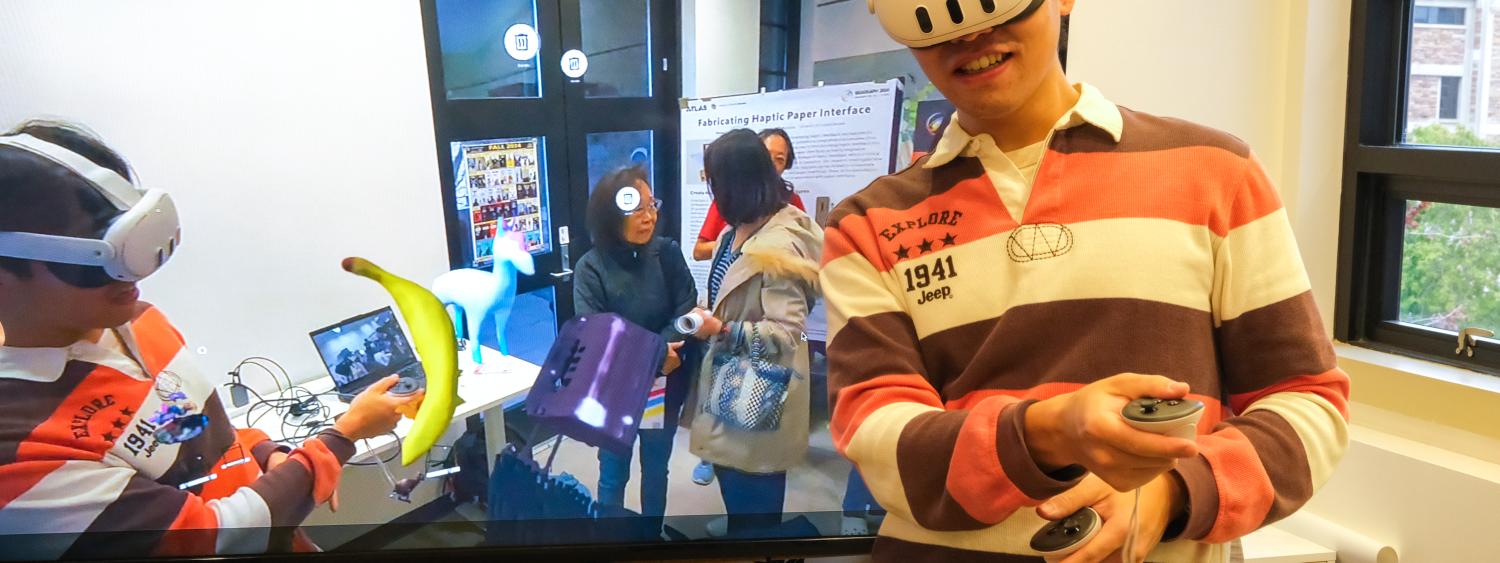 A man stands in front of a TV screen wearing a VR headset. In the TV screen, he is visible interacting with a 3d model of a banana.