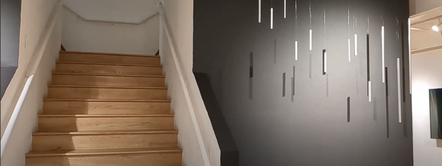 A set of stairs next to a gray wall. The gray wall is lit with a spotlight and features several metal cylinders hanging from the ceiling.