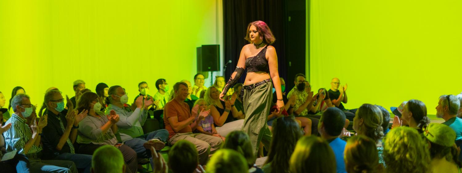 Bright lime green lighting is projected on the walls in a dark room. A model walks down the center of the screen with an audience surrounding them.