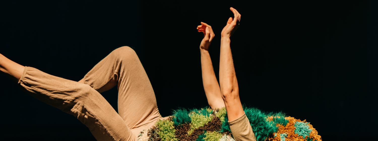 A person in a dark room lays on the floor, arms and legs up in the air. Their torso and face is covered by a rug resembling moss.