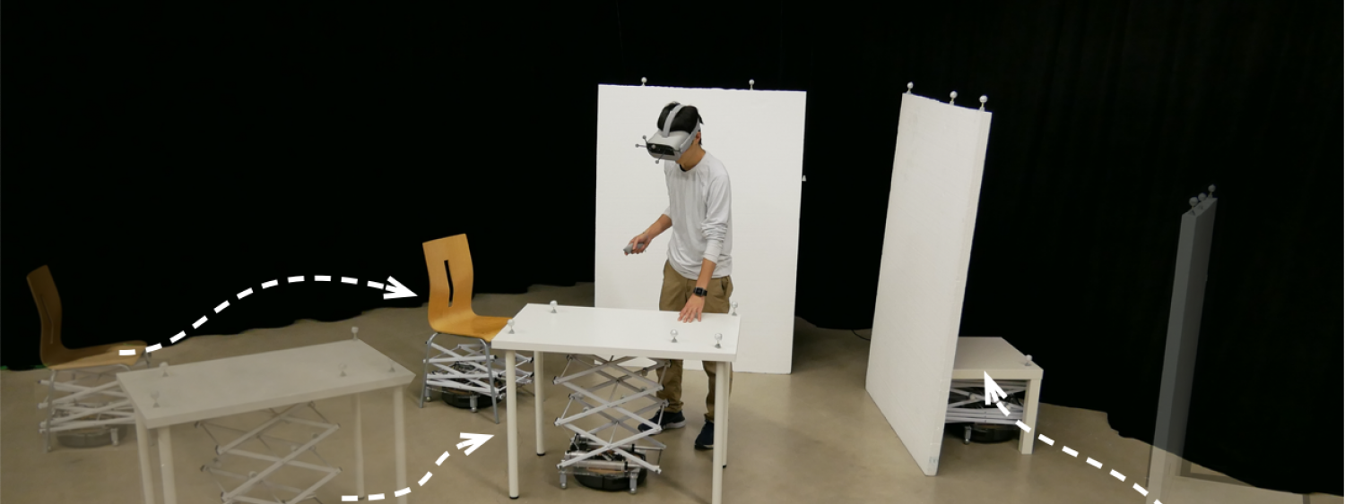 Person with VR goggles touches a table that has a Roomba with a mechanical scissor lift below.