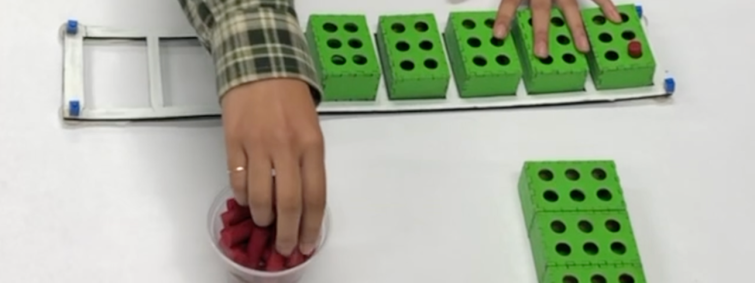 A BrailleBlocks user building a word with the blocks and pegs. There is a white frame on the table with rectangle slots. There are 8 slots in a row. 5 of the slots have green blocks in them which resemble Braille cells. The user is reaching for red pegs which she is placing in the block holes to create Braille letters.