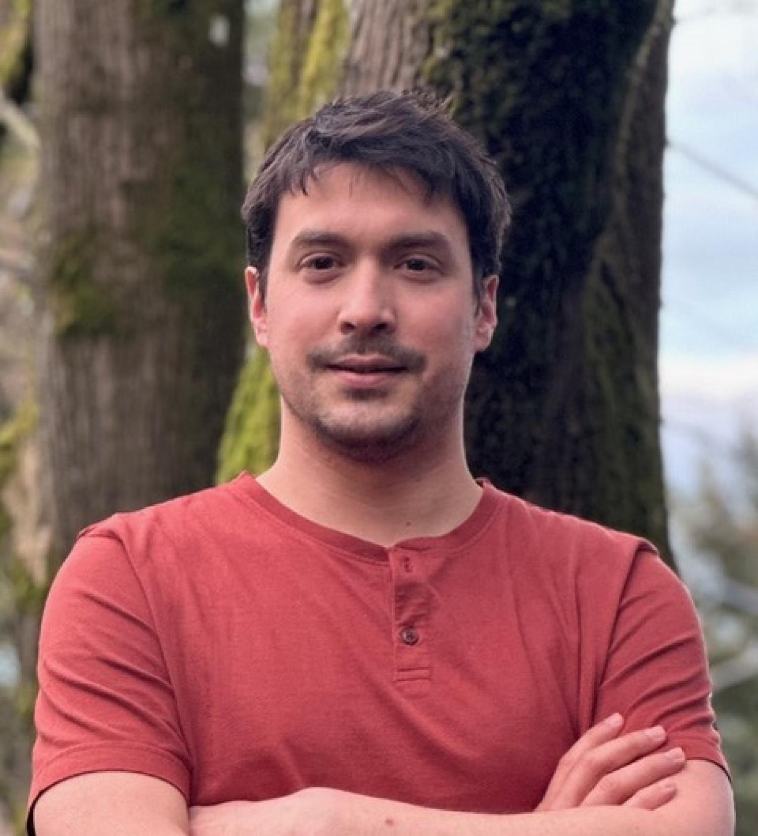 Headshot of Nathan Belliveau standing with crossed arms in front of a tree