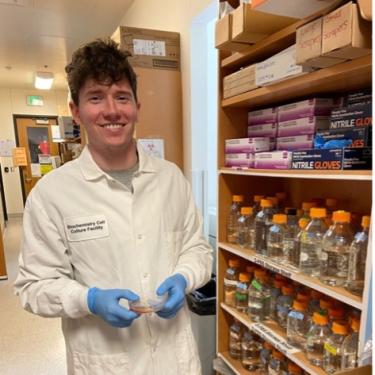 A scientist in a lab coat holding a dish of cells in front of a shelf of lab supplies.