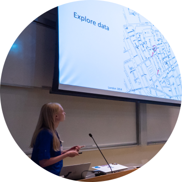 A woman at a podium looking at a screen projecting the words "Explore data"
