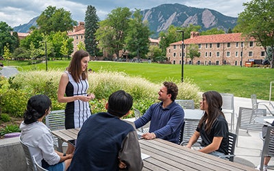 Alix leads a discussion among students seated outside the Koelbel Building. Mountains are visible in the background.