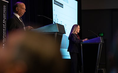 Two presenters at the front of the room, both in professional attire.