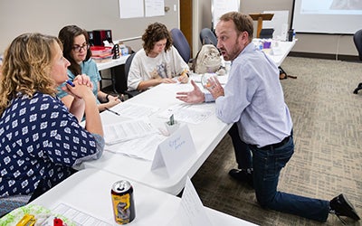 Erick Mueller kneels as he talks to entrepreneurs, seated at a table, at a founders workshop.