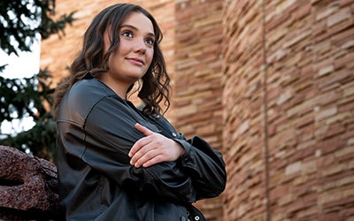 Chloe poses with her arms crossed against a rock. A sandstone campus building is visible in the background.