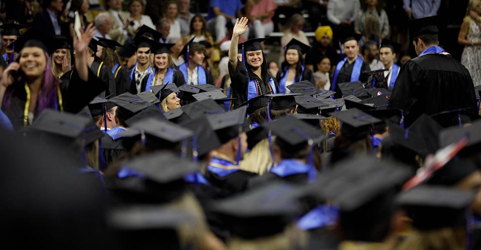 A woman in regalia waves to the camera. In the foreground are her classmates in a large stadium.