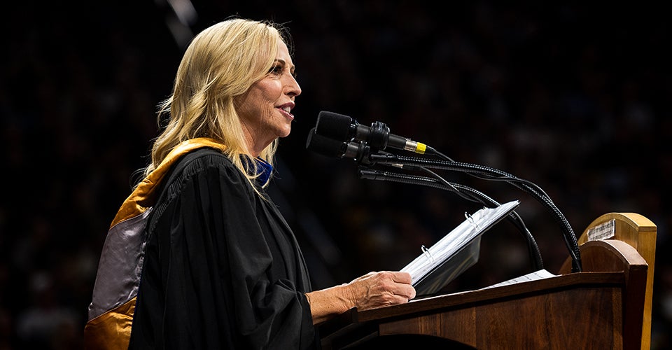 Jay Parry, in her regalia, speaks to the graduates at the podium of the CU Events Center.