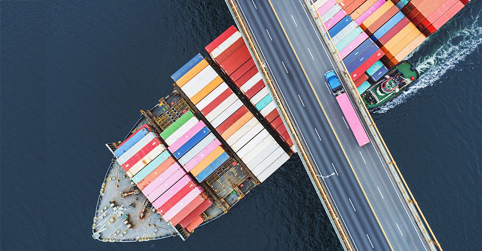 A full container ship passes under a bridge, where a lone truck is driving.