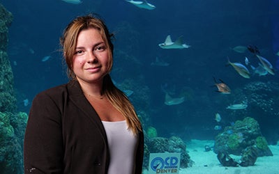 Close up of Emma Pearson standing in the Denver Aquarium as fish and rays swim by.