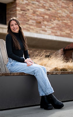 Kyla Thomas laughs as she sits on a bench outside the Koelbel Building on a sunny day.