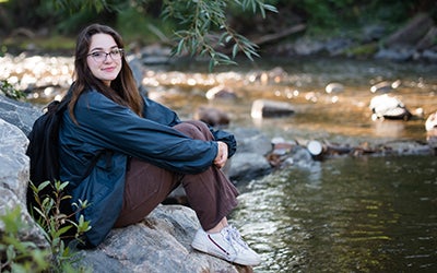 Julie Gentile smiles in an outdoor scene. She's sitting on a rock near a river.