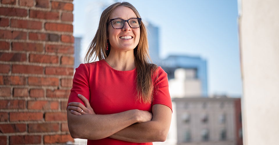 Colette, in a red dress, looks out over the Denver skyline from the roof of her building.