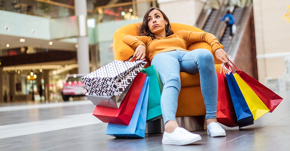 A young woman sits in a chair in a mall, looking exhausted, surrounded by small shopping bags.