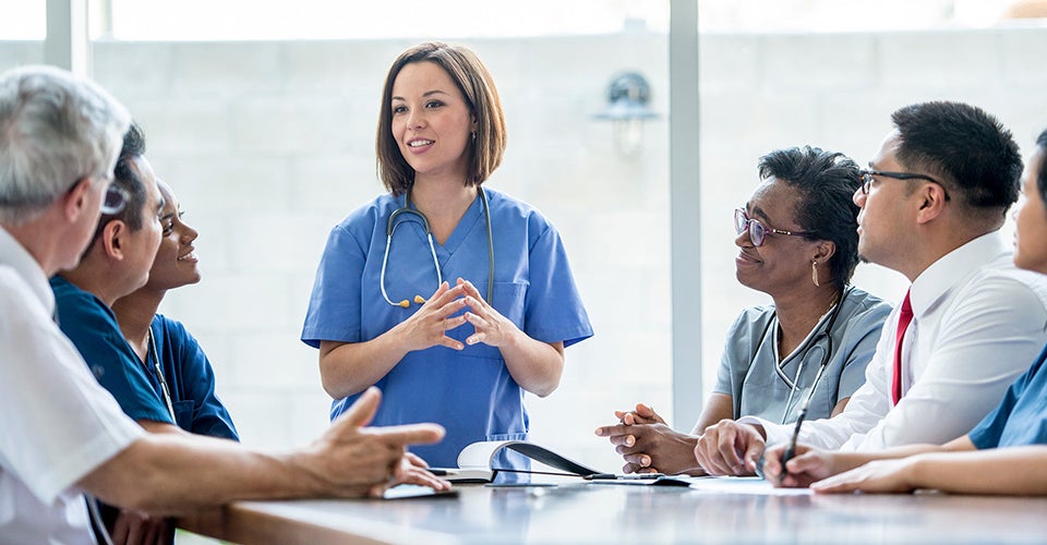 A woman in blue scrubs leads a presentation to a team of professionals in a conference room.