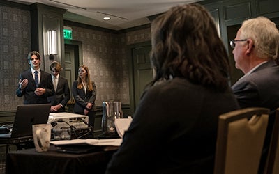 Two students present in the background as the judges watch in the foreground.